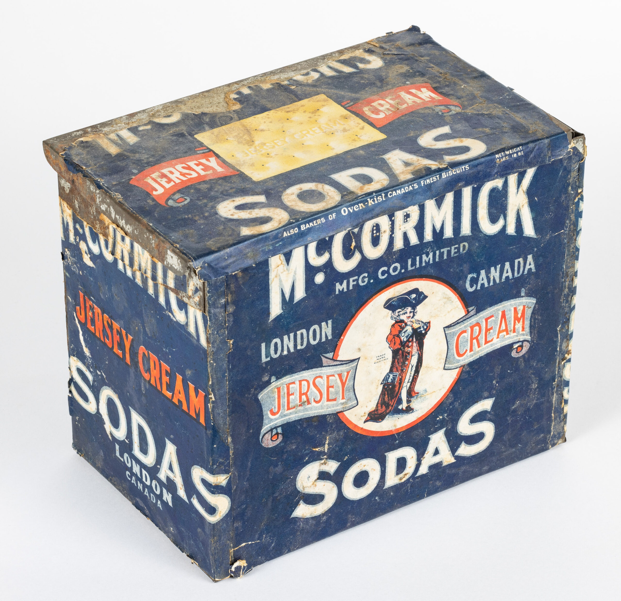 A slightly rusting tin box is revealed by the worn paper label that once fully covered it. The label is covered in lettering advertising Jersey Cream Sodas by McCormick. There is a small illustration on the front of the box of a youth in British regalia, complete with a jaunty 3-pointed hat and long red military coat that is far too large for his size.