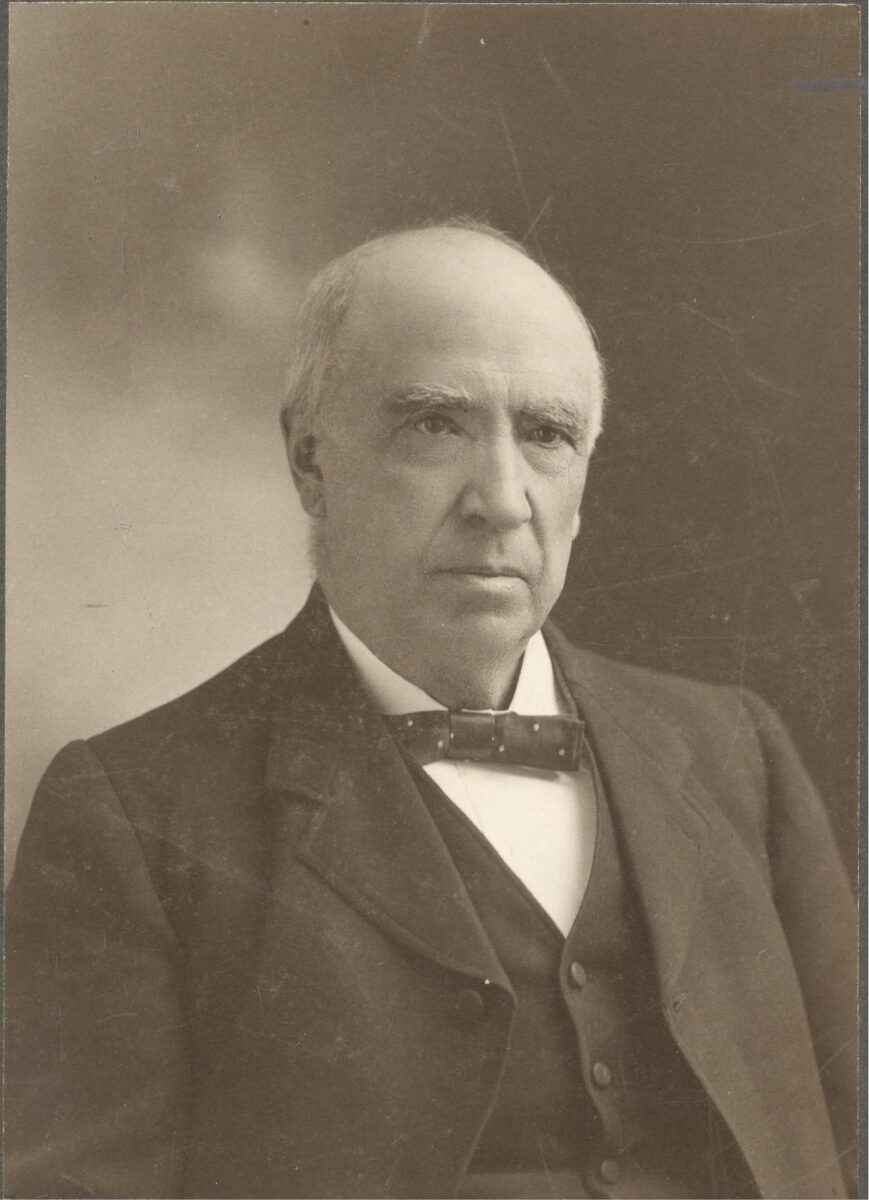 A sepia-toned photograph shows a balding man with soft features and bushy eyebrows. He wears a dotted bowtie and a neat vest and suit jacket with minimal lapels.