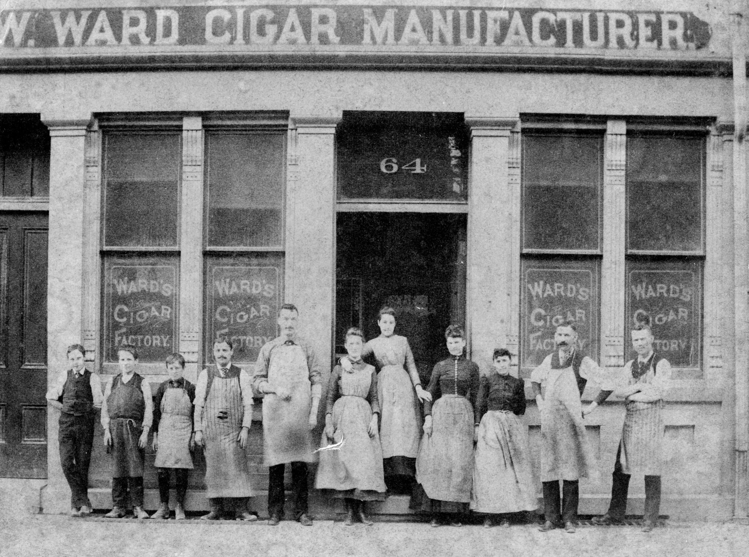 A grainy black and white photograph of the staff of W. Ward Cigar Manufacturer. 4 men with the handlebar moustaches and aprons of that era, 4 women wearing petticoats, and 3 young boys stand outside the building in a row.