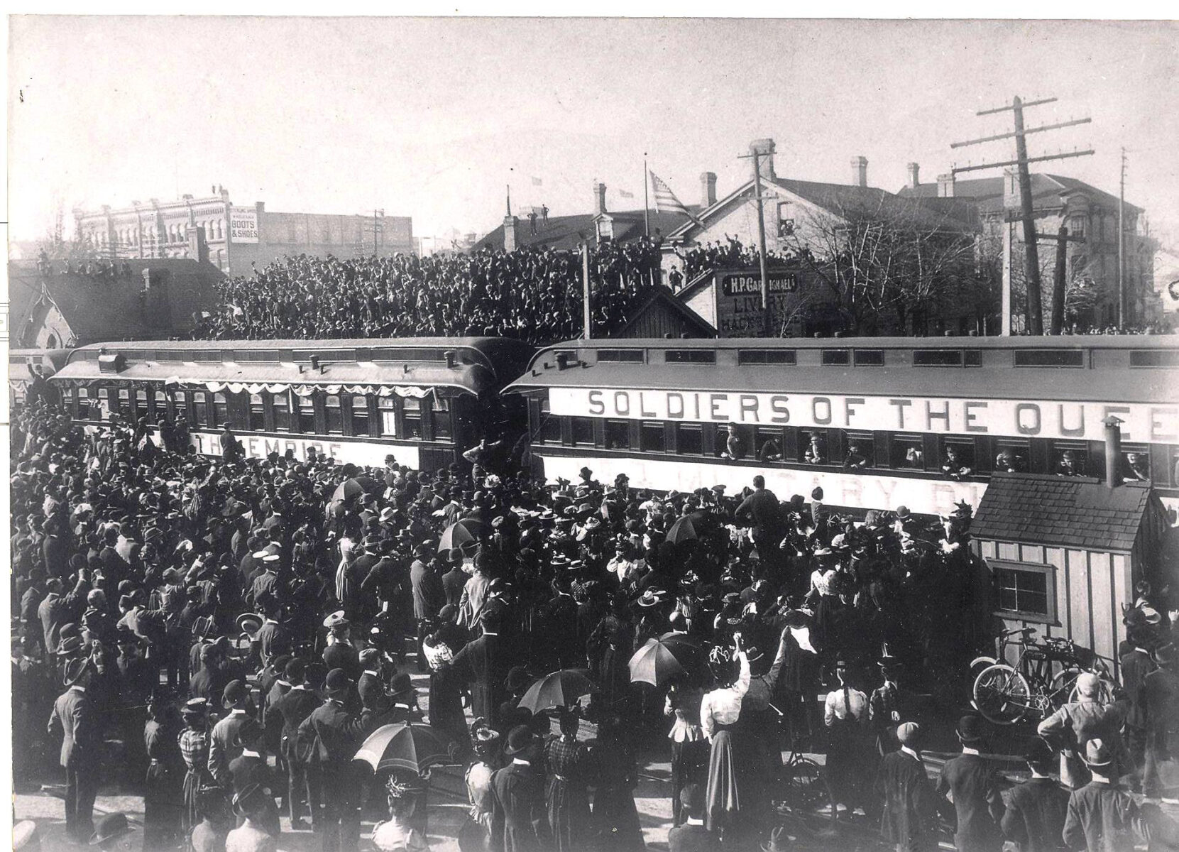 A faded black and white photograph depicts a train at a train station crowded in a sea of people. People stand on the roofs of the buildings in the distance, crowding the scene.